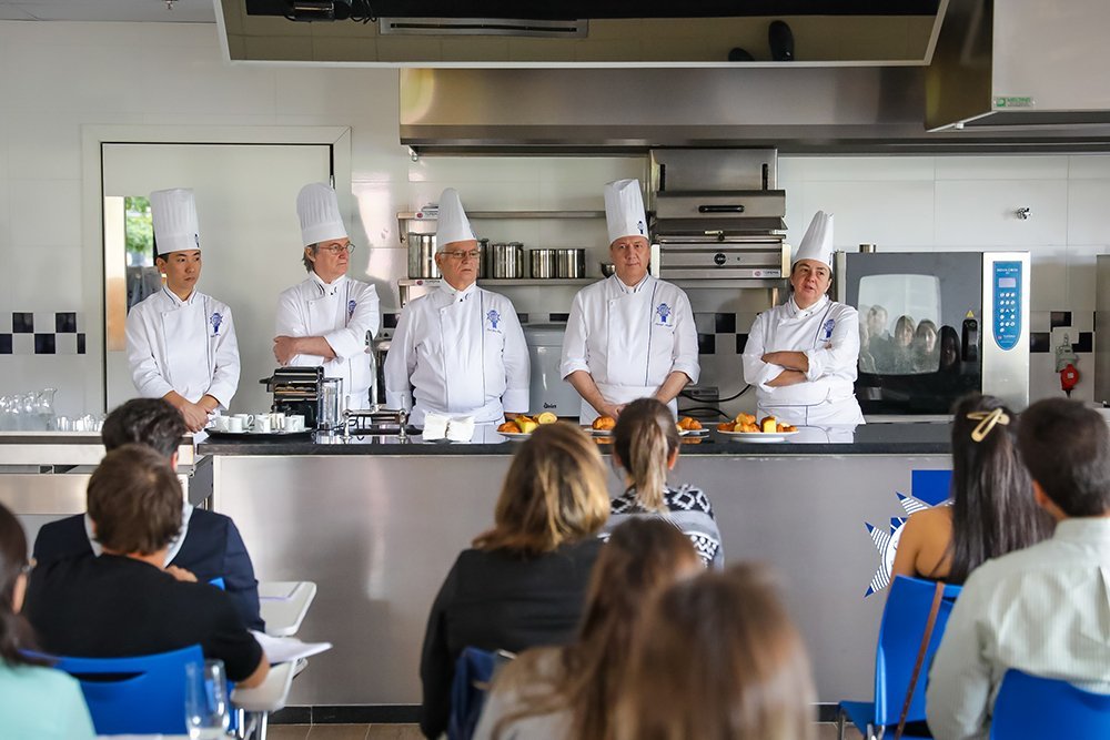 Fotografia de um aluno da Le Cordon Bleu, vestindo dólmã branca com o logo azul, focado na finalização de pratos em uma cozinha profissional de ensino.